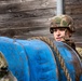 A soldier in camouflage uniform and helmet is handling a large blue barrel or drum covered in mud and rope. The soldier appears to be participating in a military training exercise, likely an obstacle course or physical training drill. A wooden plank wall serves as the background.