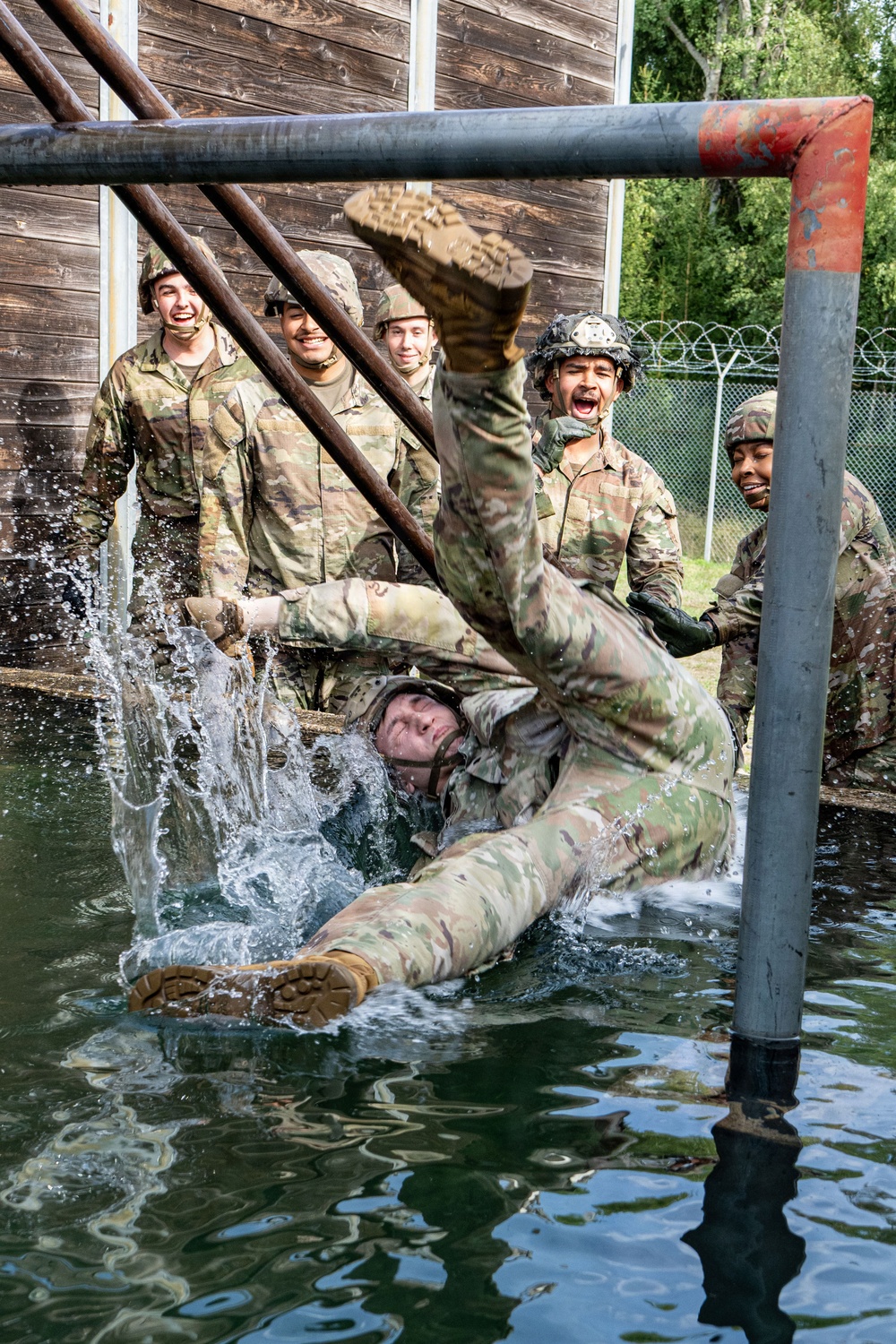 A group of military personnel in camouflage uniforms and helmets are participating in a water obstacle course training exercise. One soldier is falling or being dunked into a water pit while teammates watch and react with laughter and excitement. The scene takes place at what appears to be a military training facility with wooden structures, metal pipe obstacles, and a chain-link fence with razor wire in the background.