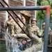 A group of military personnel in camouflage uniforms and helmets are participating in a water obstacle course training exercise. One soldier is falling or being dunked into a water pit while teammates watch and react with laughter and excitement. The scene takes place at what appears to be a military training facility with wooden structures, metal pipe obstacles, and a chain-link fence with razor wire in the background.