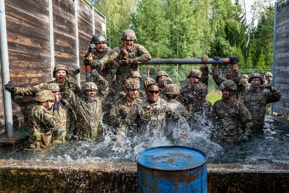 A large group of military personnel in full combat gear and helmets are submerged or standing in a water obstacle during what appears to be a military training exercise. The soldiers are visibly energized and shouting, with water splashing around them. The setting includes wooden walls, a chain-link fence, and green trees in the background, consistent with a military training facility.