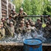 A large group of military personnel in full combat gear and helmets are submerged or standing in a water obstacle during what appears to be a military training exercise. The soldiers are visibly energized and shouting, with water splashing around them. The setting includes wooden walls, a chain-link fence, and green trees in the background, consistent with a military training facility.