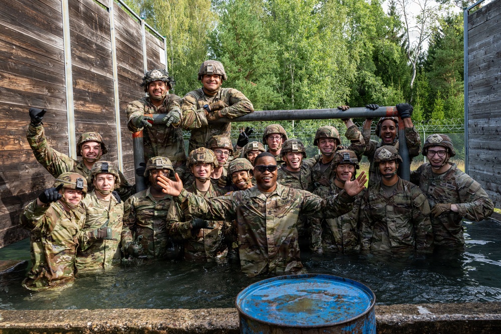 A large group of military personnel in camouflage uniforms and helmets are posing together in a water obstacle pit, likely during a training exercise. The soldiers appear to be in high spirits, smiling and waving at the camera after completing what appears to be a water obstacle course challenge. The setting includes wooden barrier walls, a chain-link fence, and surrounding trees consistent with a military training facility.