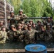 A large group of military personnel in camouflage uniforms and helmets are posing together in a water obstacle pit, likely during a training exercise. The soldiers appear to be in high spirits, smiling and waving at the camera after completing what appears to be a water obstacle course challenge. The setting includes wooden barrier walls, a chain-link fence, and surrounding trees consistent with a military training facility.