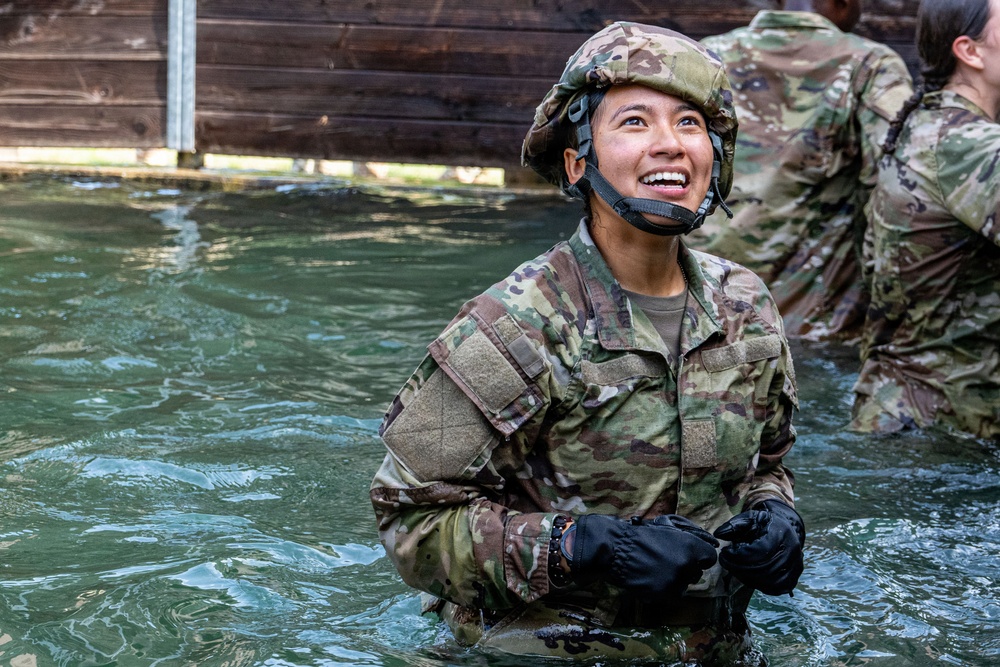 A soldier in full camouflage uniform and helmet is standing waist-deep in water, smiling broadly and looking upward. The individual is wearing black tactical gloves and a chin-strapped helmet, suggesting participation in a military water obstacle or training exercise. At least one other soldier in similar gear is visible in the background.