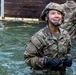 A soldier in full camouflage uniform and helmet is standing waist-deep in water, smiling broadly and looking upward. The individual is wearing black tactical gloves and a chin-strapped helmet, suggesting participation in a military water obstacle or training exercise. At least one other soldier in similar gear is visible in the background.