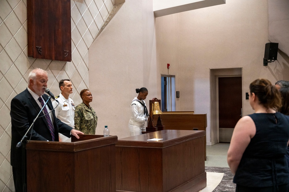 A formal military ceremony is taking place in what appears to be a chapel or ceremonial hall. A civilian man in a dark suit speaks at a podium/lectern while two Navy personnel in uniform stand at attention nearby, and a sailor in dress whites stands beside a ceremonial brass bell. A woman in civilian attire observes from the right foreground.