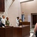A formal military ceremony is taking place in what appears to be a chapel or ceremonial hall. A civilian man in a dark suit speaks at a podium/lectern while two Navy personnel in uniform stand at attention nearby, and a sailor in dress whites stands beside a ceremonial brass bell. A woman in civilian attire observes from the right foreground.