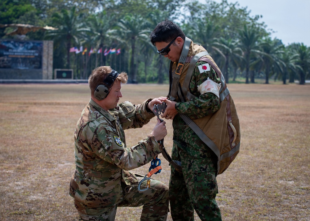 Two military personnel are engaged in a pre-jump equipment check on an open field. One soldier, wearing U.S. Army OCP (Operational Camouflage Pattern) uniform with hearing protection, is kneeling and assisting the other with parachute or rappelling harness equipment. The standing soldier wears Japanese Ground Self-Defense Force (JGSDF) camouflage uniform with a visible Japanese flag patch, suggesting a multinational military exercise.