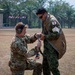 Two military personnel are engaged in a pre-jump equipment check on an open field. One soldier, wearing U.S. Army OCP (Operational Camouflage Pattern) uniform with hearing protection, is kneeling and assisting the other with parachute or rappelling harness equipment. The standing soldier wears Japanese Ground Self-Defense Force (JGSDF) camouflage uniform with a visible Japanese flag patch, suggesting a multinational military exercise.