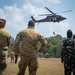 Military personnel from multiple nations observe a medical evacuation (MEDEVAC) helicopter hoist operation on an open field. A UH-60 Black Hawk helicopter marked with a Red Cross symbol is performing a hoist extraction, lifting what appears to be a person or litter. The scene appears to be a joint multinational military training exercise.