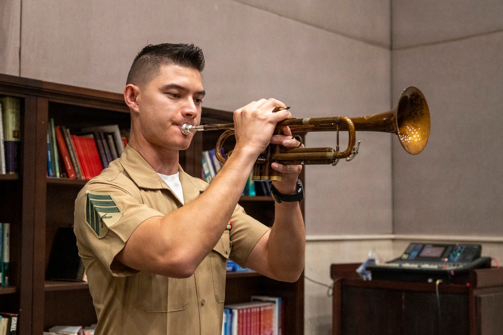 A young male in a military uniform is playing a brass trumpet in what appears to be an indoor office or library setting. The instrument shows signs of age and wear, with a patinated, golden-brown finish. The subject appears focused and engaged in active performance.
