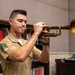 A young male in a military uniform is playing a brass trumpet in what appears to be an indoor office or library setting. The instrument shows signs of age and wear, with a patinated, golden-brown finish. The subject appears focused and engaged in active performance.