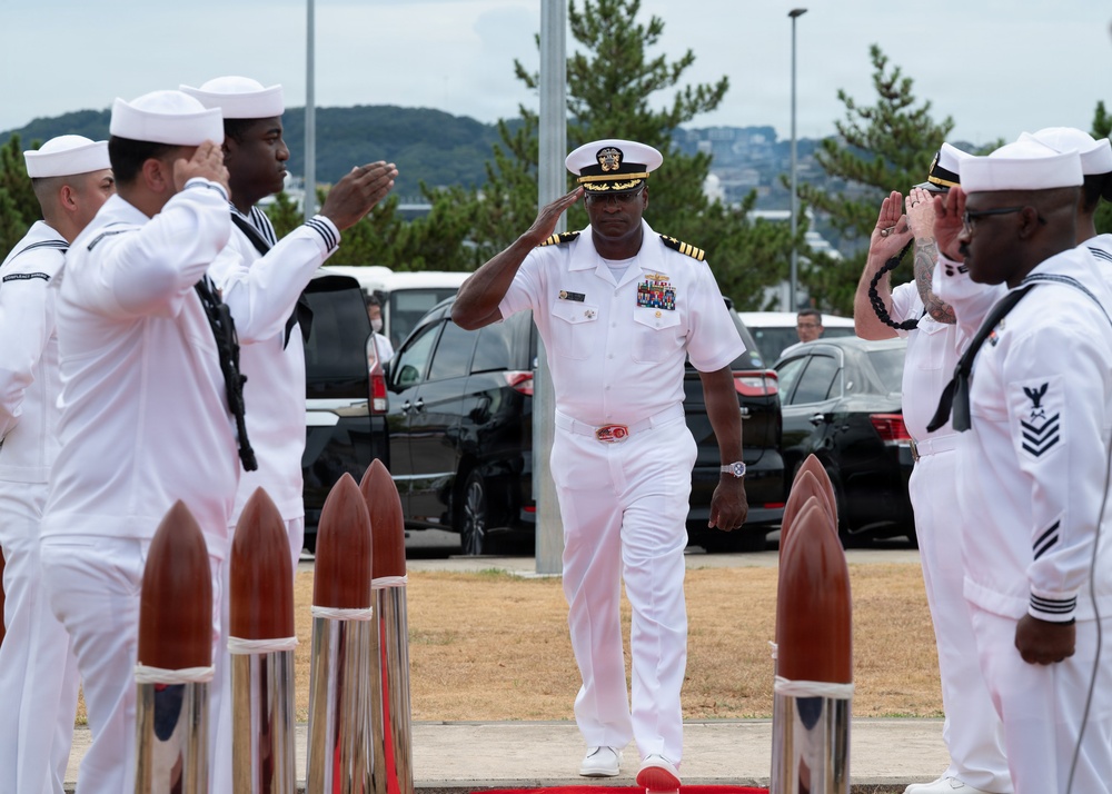 A senior U.S. Navy officer in white dress uniform walks between two rows of enlisted sailors who are rendering a salute in a formal sideboy ceremony. The officer, wearing a combination cover with gold braid and multiple service ribbons, is returning the salute while walking along a red carpet flanked by decorative bollards. The setting appears to be an outdoor naval installation, likely in Japan based on the visible landscape.