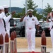 A senior U.S. Navy officer in white dress uniform walks between two rows of enlisted sailors who are rendering a salute in a formal sideboy ceremony. The officer, wearing a combination cover with gold braid and multiple service ribbons, is returning the salute while walking along a red carpet flanked by decorative bollards. The setting appears to be an outdoor naval installation, likely in Japan based on the visible landscape.