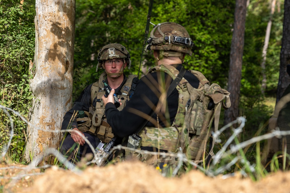 Two military personnel in full tactical gear are positioned near a tree in a forested environment, appearing to communicate or receive a briefing. One soldier faces the camera wearing glasses and a helmet with a mounted device, while the other has their back turned and carries a radio antenna and rucksack. Barbed wire is visible in the foreground, suggesting a defensive perimeter or training exercise.
