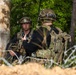 Two military personnel in full tactical gear are positioned near a tree in a forested environment, appearing to communicate or receive a briefing. One soldier faces the camera wearing glasses and a helmet with a mounted device, while the other has their back turned and carries a radio antenna and rucksack. Barbed wire is visible in the foreground, suggesting a defensive perimeter or training exercise.