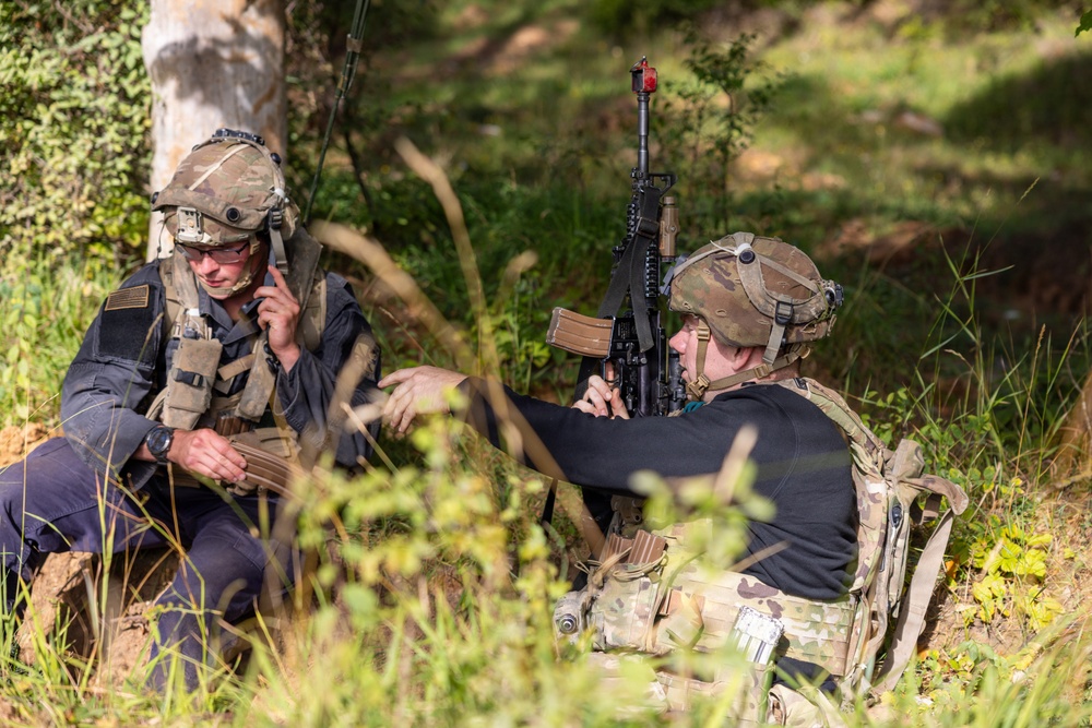 Two armed individuals in tactical military gear are positioned in a wooded outdoor area. One person is using a radio/communication device while the other holds an AK-pattern rifle in a low-profile position. Both wear camouflage helmets with mounted accessories and tactical vests.