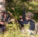 Two armed individuals in tactical military gear are positioned in a wooded outdoor area. One person is using a radio/communication device while the other holds an AK-pattern rifle in a low-profile position. Both wear camouflage helmets with mounted accessories and tactical vests.