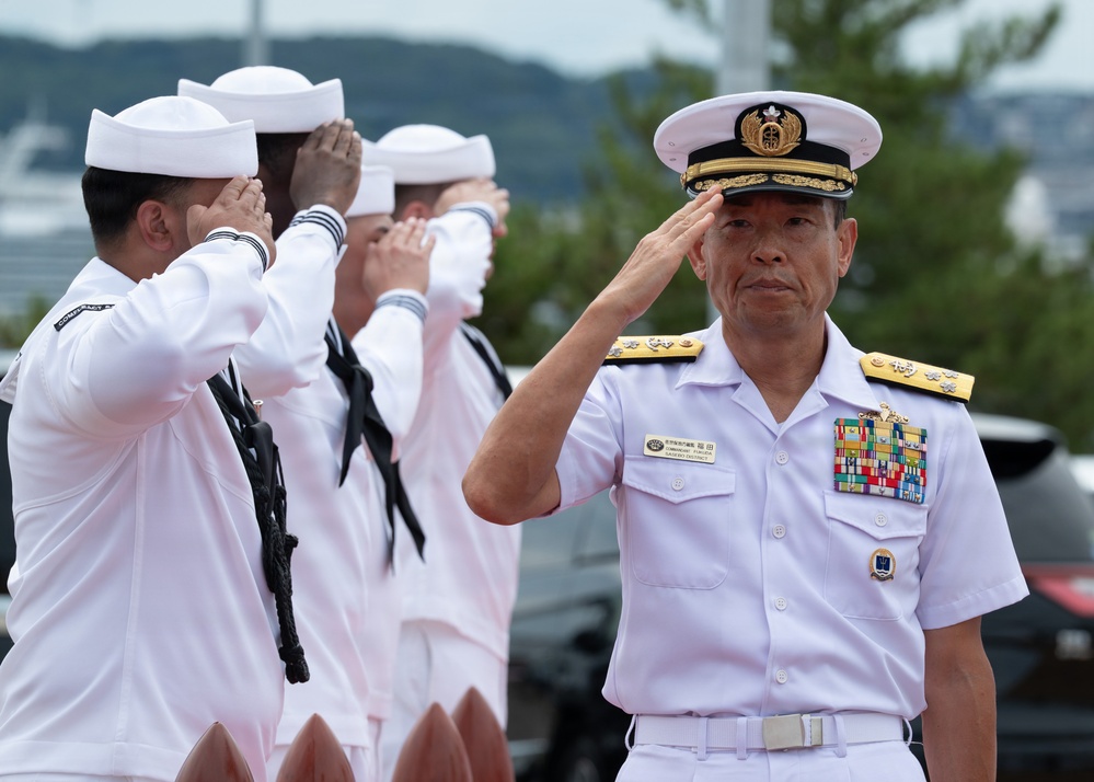 A senior naval officer in a white dress uniform with gold epaulettes and multiple service ribbons renders a salute during what appears to be a formal military ceremony. Behind him, several sailors in white enlisted uniforms also salute in unison. The setting appears to be an outdoor naval installation, likely in Japan based on the officer's uniform insignia and name tag.