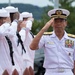 A senior naval officer in a white dress uniform with gold epaulettes and multiple service ribbons renders a salute during what appears to be a formal military ceremony. Behind him, several sailors in white enlisted uniforms also salute in unison. The setting appears to be an outdoor naval installation, likely in Japan based on the officer's uniform insignia and name tag.