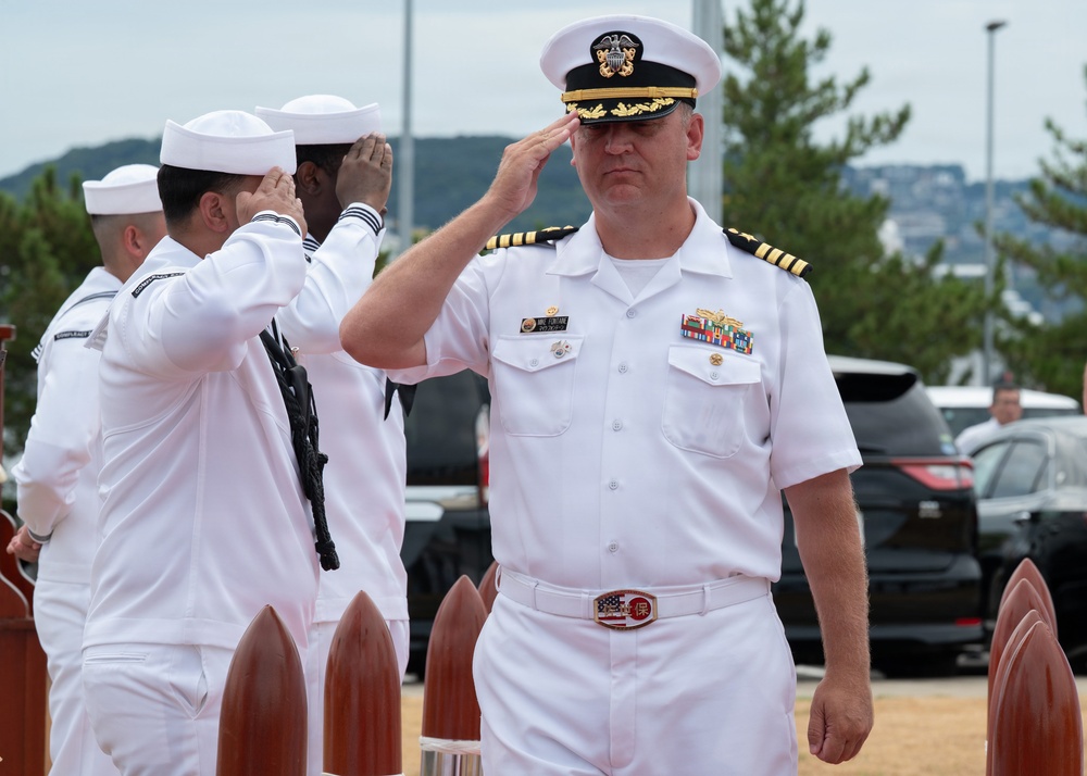 A U.S. Navy Captain (O-6) in white service dress uniform renders a salute during what appears to be a formal military ceremony. Several enlisted sailors in white uniforms are also saluting in the background. The setting appears to be an outdoor naval installation with decorative wooden bollards visible in the foreground.