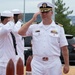 A U.S. Navy Captain (O-6) in white service dress uniform renders a salute during what appears to be a formal military ceremony. Several enlisted sailors in white uniforms are also saluting in the background. The setting appears to be an outdoor naval installation with decorative wooden bollards visible in the foreground.