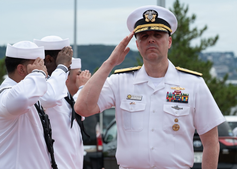 A senior U.S. Navy officer in white dress uniform renders a salute during what appears to be a formal military ceremony. Behind him, enlisted sailors in white service uniforms also salute, suggesting a ceremonial event such as a change of command, arrival honors, or official review. The setting appears to be an outdoor military installation.