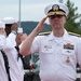 A senior U.S. Navy officer in white dress uniform renders a salute during what appears to be a formal military ceremony. Behind him, enlisted sailors in white service uniforms also salute, suggesting a ceremonial event such as a change of command, arrival honors, or official review. The setting appears to be an outdoor military installation.