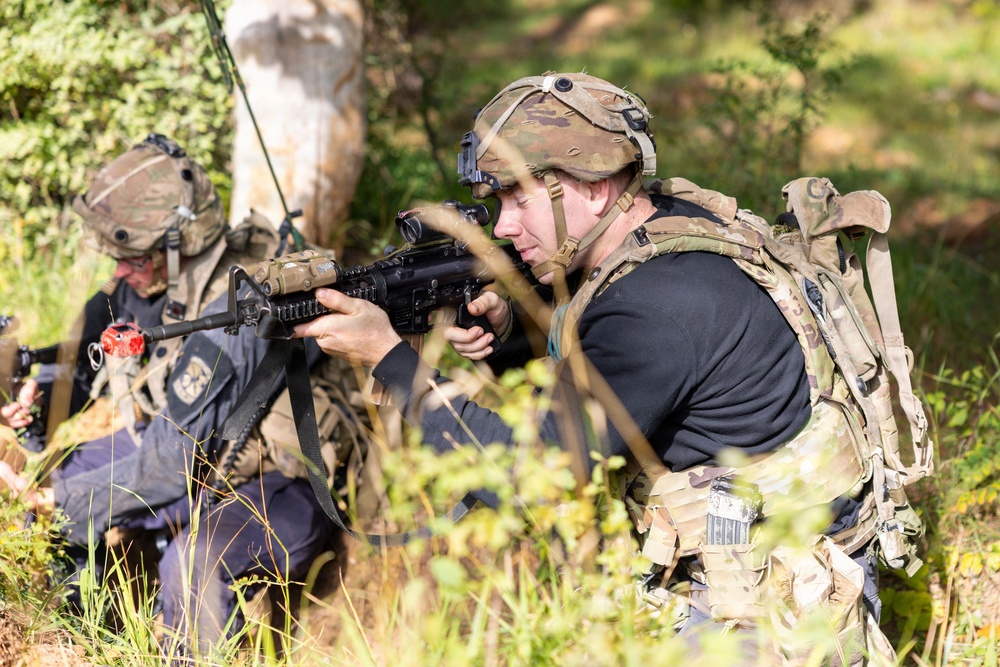 Two individuals in military-style tactical gear are positioned in a wooded outdoor environment. The foreground figure is aiming a rifle with an optical sight while wearing a combat helmet, plate carrier vest, and rucksack. A second figure in the background appears to be crouching near a tree, also in full tactical kit.