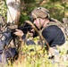 Two individuals in military-style tactical gear are positioned in a wooded outdoor environment. The foreground figure is aiming a rifle with an optical sight while wearing a combat helmet, plate carrier vest, and rucksack. A second figure in the background appears to be crouching near a tree, also in full tactical kit.