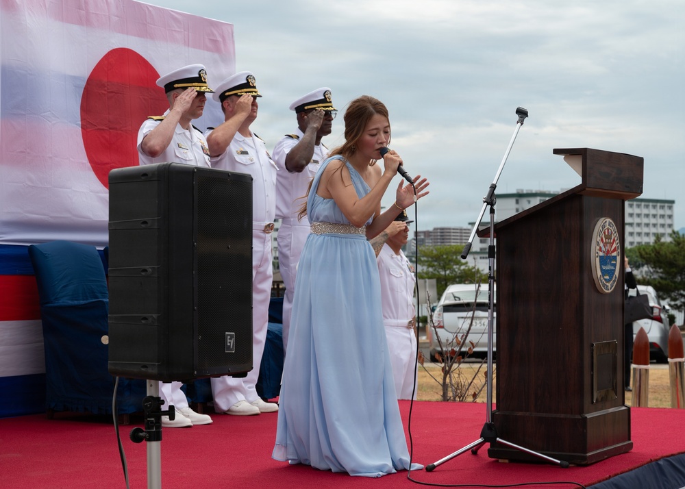 A formal military ceremony is taking place on an outdoor stage with a red carpet. A young woman in a light blue evening gown is singing into a microphone while U.S. Navy officers in white dress uniforms stand at salute behind her. A Japanese flag and what appears to be a U.S. flag are visible in the background, suggesting a bilateral military event.