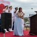 A formal military ceremony is taking place on an outdoor stage with a red carpet. A young woman in a light blue evening gown is singing into a microphone while U.S. Navy officers in white dress uniforms stand at salute behind her. A Japanese flag and what appears to be a U.S. flag are visible in the background, suggesting a bilateral military event.