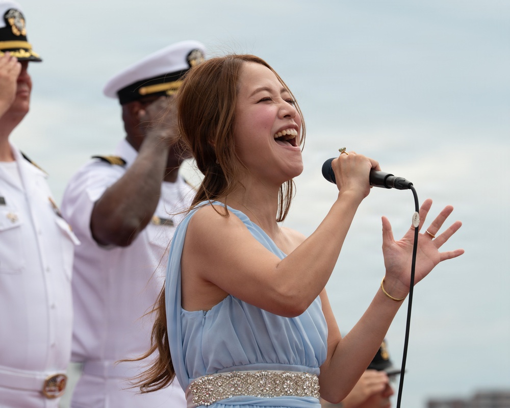 A woman in a light blue dress is singing passionately into a handheld microphone at what appears to be an outdoor military ceremony or event. She is performing in front of uniformed naval officers dressed in white dress uniforms. Her expression conveys strong emotional engagement with the performance.