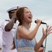 A woman in a light blue dress is singing passionately into a handheld microphone at what appears to be an outdoor military ceremony or event. She is performing in front of uniformed naval officers dressed in white dress uniforms. Her expression conveys strong emotional engagement with the performance.