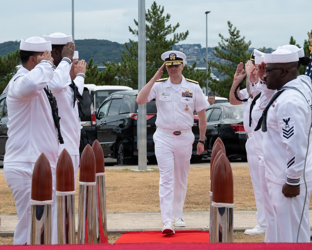 A U.S. Navy officer in white dress uniform walks along a red carpet while being saluted by enlisted sailors on both sides in a formal sideboy ceremony. The officer, wearing a combination cover with gold insignia and multiple ribbons/medals, returns the salute while passing between decorative wooden and chrome bollards. The setting appears to be an outdoor naval installation, likely in Japan based on the surrounding landscape.