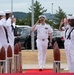 A U.S. Navy officer in white dress uniform walks along a red carpet while being saluted by enlisted sailors on both sides in a formal sideboy ceremony. The officer, wearing a combination cover with gold insignia and multiple ribbons/medals, returns the salute while passing between decorative wooden and chrome bollards. The setting appears to be an outdoor naval installation, likely in Japan based on the surrounding landscape.