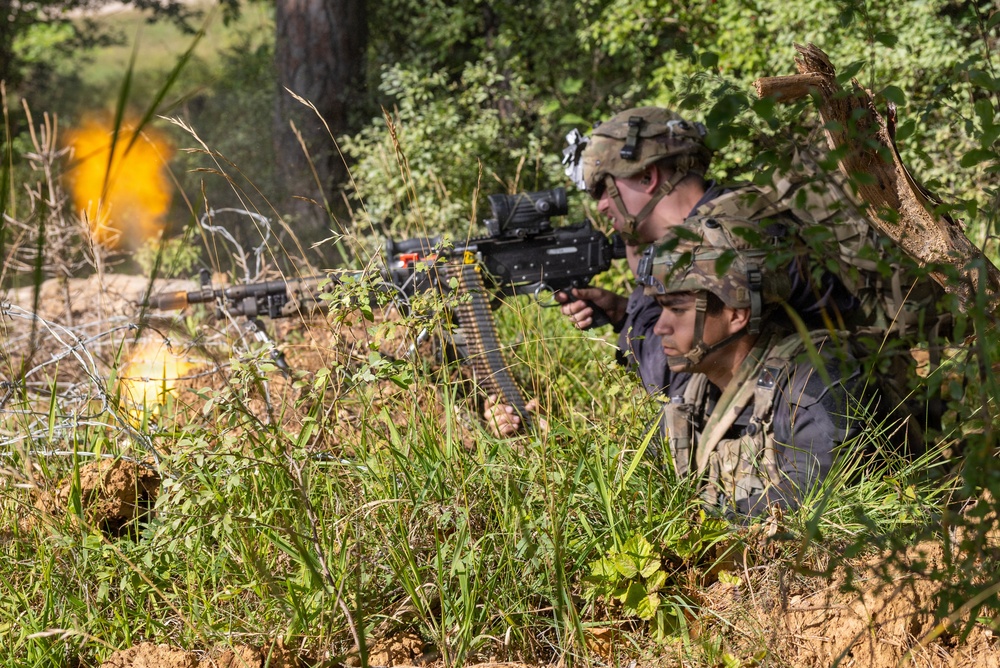 Two soldiers in full combat gear are positioned in a prone/crouched defensive position at a treeline, with one actively firing a belt-fed machine gun. Muzzle flash and an explosion or pyrotechnic effect are visible to the left, suggesting a live-fire training exercise or combat scenario. The setting is a wooded, vegetated area with visible concertina wire in the background.