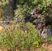 Two soldiers in full combat gear are positioned in a prone/crouched defensive position at a treeline, with one actively firing a belt-fed machine gun. Muzzle flash and an explosion or pyrotechnic effect are visible to the left, suggesting a live-fire training exercise or combat scenario. The setting is a wooded, vegetated area with visible concertina wire in the background.