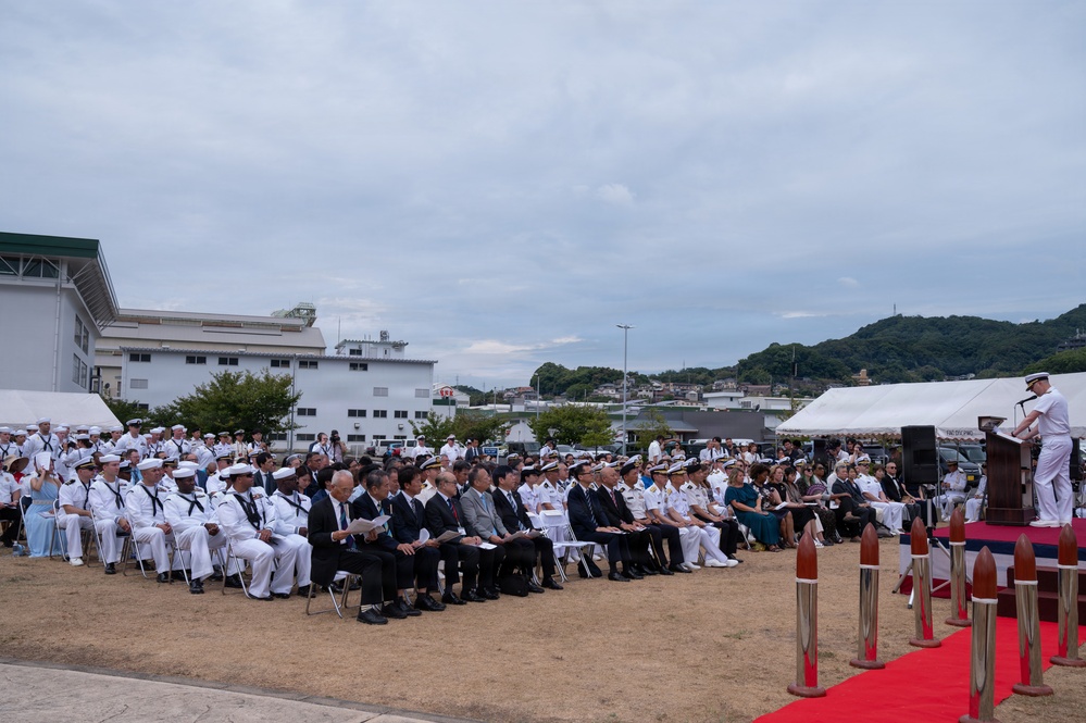 A formal military ceremony is taking place outdoors at what appears to be a naval installation, likely in Japan based on the architectural style and mixed attendance of U.S. Navy personnel and Japanese civilians and military officers. A speaker in white naval dress uniform addresses a large seated audience from a podium on a red carpet. The crowd includes both American and Japanese naval officers, civilian dignitaries, and other attendees.