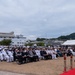 A formal military ceremony is taking place outdoors at what appears to be a naval installation, likely in Japan based on the architectural style and mixed attendance of U.S. Navy personnel and Japanese civilians and military officers. A speaker in white naval dress uniform addresses a large seated audience from a podium on a red carpet. The crowd includes both American and Japanese naval officers, civilian dignitaries, and other attendees.