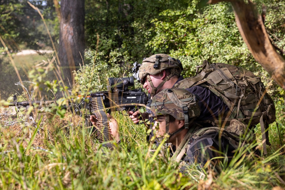 Two soldiers in full combat gear are positioned prone in dense vegetation, with one aiming a belt-fed machine gun through an optic sight. They are wearing multicam/camouflage uniforms with tactical vests and helmets equipped with night vision mounts. The setting appears to be a wooded training or operational environment during daylight hours.
