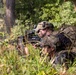 Two soldiers in full combat gear are positioned prone in dense vegetation, with one aiming a belt-fed machine gun through an optic sight. They are wearing multicam/camouflage uniforms with tactical vests and helmets equipped with night vision mounts. The setting appears to be a wooded training or operational environment during daylight hours.