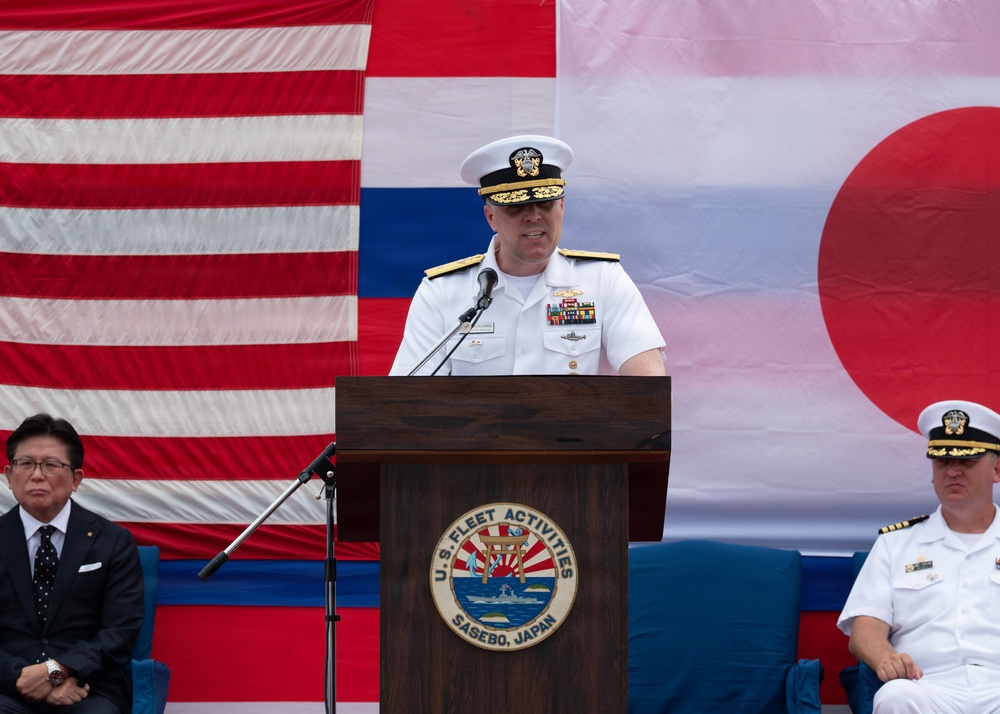 A U.S. Navy officer in white dress uniform delivers a speech at a podium bearing the seal of U.S. Fleet Activities Sasebo, Japan. The backdrop features large American and Japanese flags, indicating a formal bilateral military ceremony. Two other individuals are seated behind the speaker — one in civilian attire and one in U.S. Navy white dress uniform.