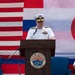A U.S. Navy officer in white dress uniform delivers a speech at a podium bearing the seal of U.S. Fleet Activities Sasebo, Japan. The backdrop features large American and Japanese flags, indicating a formal bilateral military ceremony. Two other individuals are seated behind the speaker — one in civilian attire and one in U.S. Navy white dress uniform.