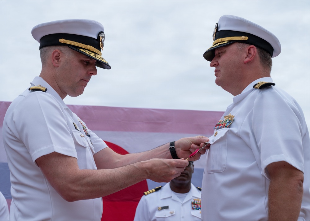 Two U.S. Navy officers in white service dress uniforms are engaged in a medal pinning ceremony, with the officer on the left attaching a decoration to the uniform of the officer on the right. A third officer in white uniform is visible in the background. The setting appears to be an outdoor military ceremony with a large flag or banner visible behind them.
