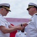 Two U.S. Navy officers in white service dress uniforms are engaged in a medal pinning ceremony, with the officer on the left attaching a decoration to the uniform of the officer on the right. A third officer in white uniform is visible in the background. The setting appears to be an outdoor military ceremony with a large flag or banner visible behind them.