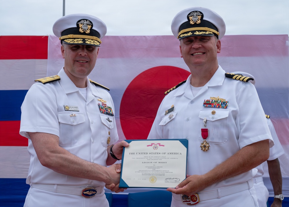 Two U.S. Navy officers in white dress uniforms are jointly holding a Legion of Merit certificate issued by the United States of America. The ceremony appears to be an awards presentation, likely a change of command or retirement ceremony, taking place outdoors with flags visible in the background. One officer is presenting the award to the other, who is already wearing the Legion of Merit medal on his uniform.