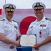 Two U.S. Navy officers in white dress uniforms are jointly holding a Legion of Merit certificate issued by the United States of America. The ceremony appears to be an awards presentation, likely a change of command or retirement ceremony, taking place outdoors with flags visible in the background. One officer is presenting the award to the other, who is already wearing the Legion of Merit medal on his uniform.