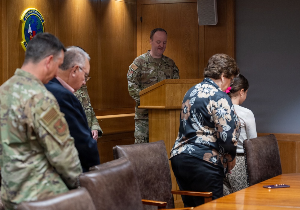 A U.S. Air Force service member in OCP (Operational Camouflage Pattern) uniform stands at a wooden podium addressing a small group of attendees in what appears to be a formal conference or briefing room. The gathering includes both military personnel in uniform and civilians, with heads bowed suggesting a moment of prayer or solemn reflection. A military unit emblem/patch is visible on the wood-paneled wall behind the speaker.