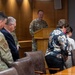 A U.S. Air Force service member in OCP (Operational Camouflage Pattern) uniform stands at a wooden podium addressing a small group of attendees in what appears to be a formal conference or briefing room. The gathering includes both military personnel in uniform and civilians, with heads bowed suggesting a moment of prayer or solemn reflection. A military unit emblem/patch is visible on the wood-paneled wall behind the speaker.